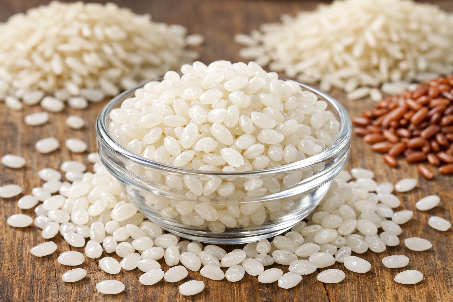 Close-up of sushi rice grains in a small bowl on a wooden table with other rice varieties blurred in the background.