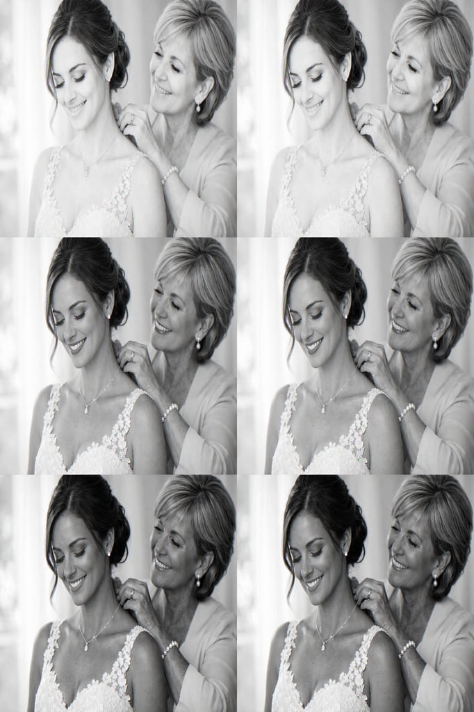 Mother fastening a necklace on her daughter on the daughter's wedding day, both smiling warmly.