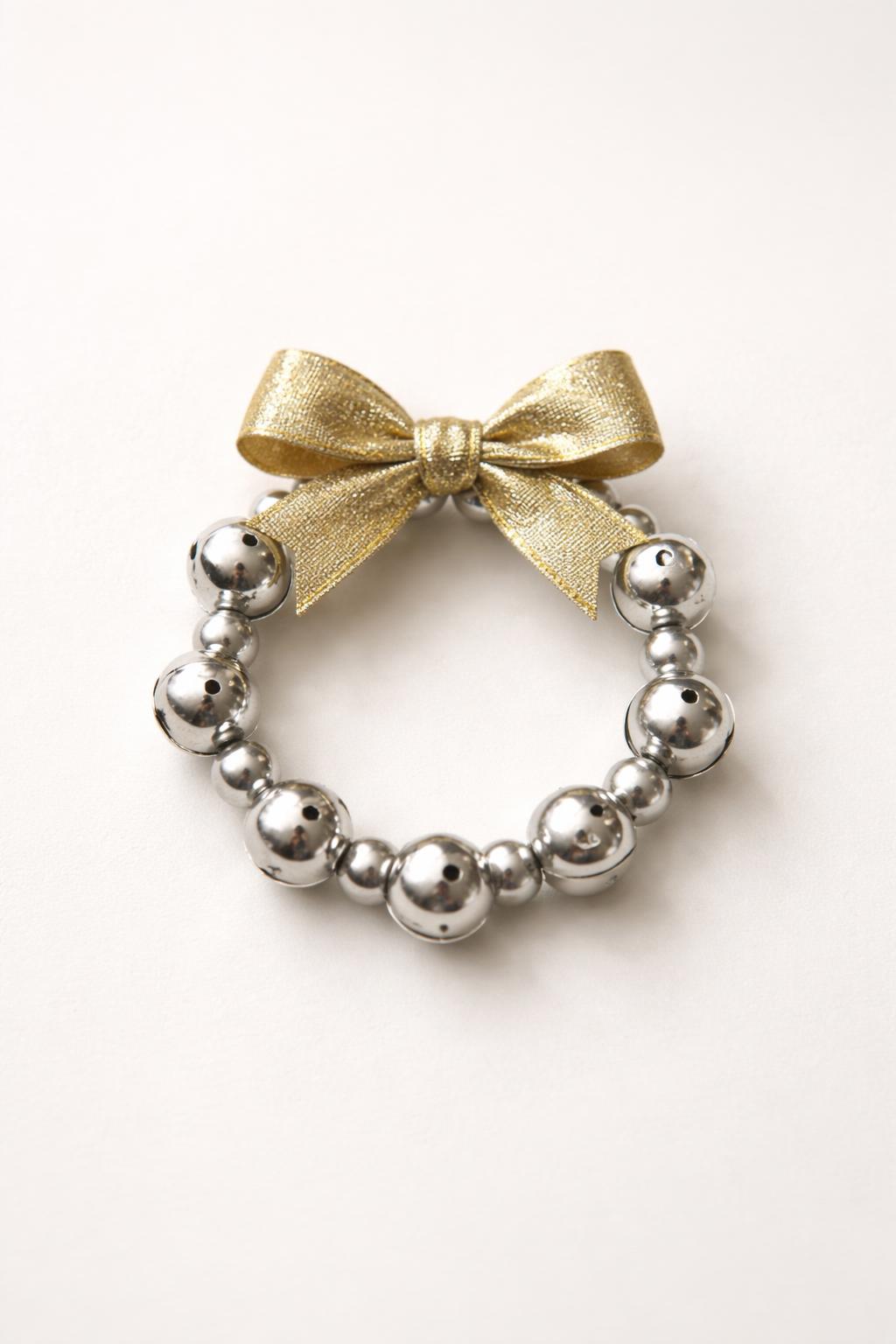 A small metallic mini wreath decorated with a gold ribbon and silver jingle bells on a plain background.