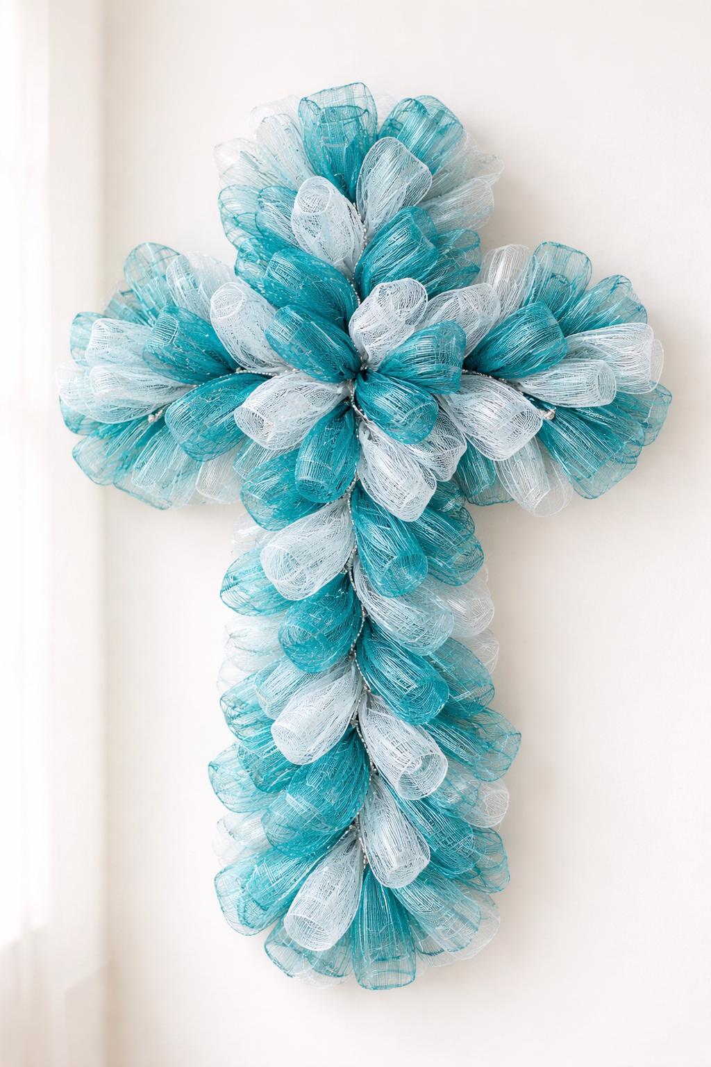 A decorative cross-shaped wreath made with teal and white mesh material displayed against a plain background.