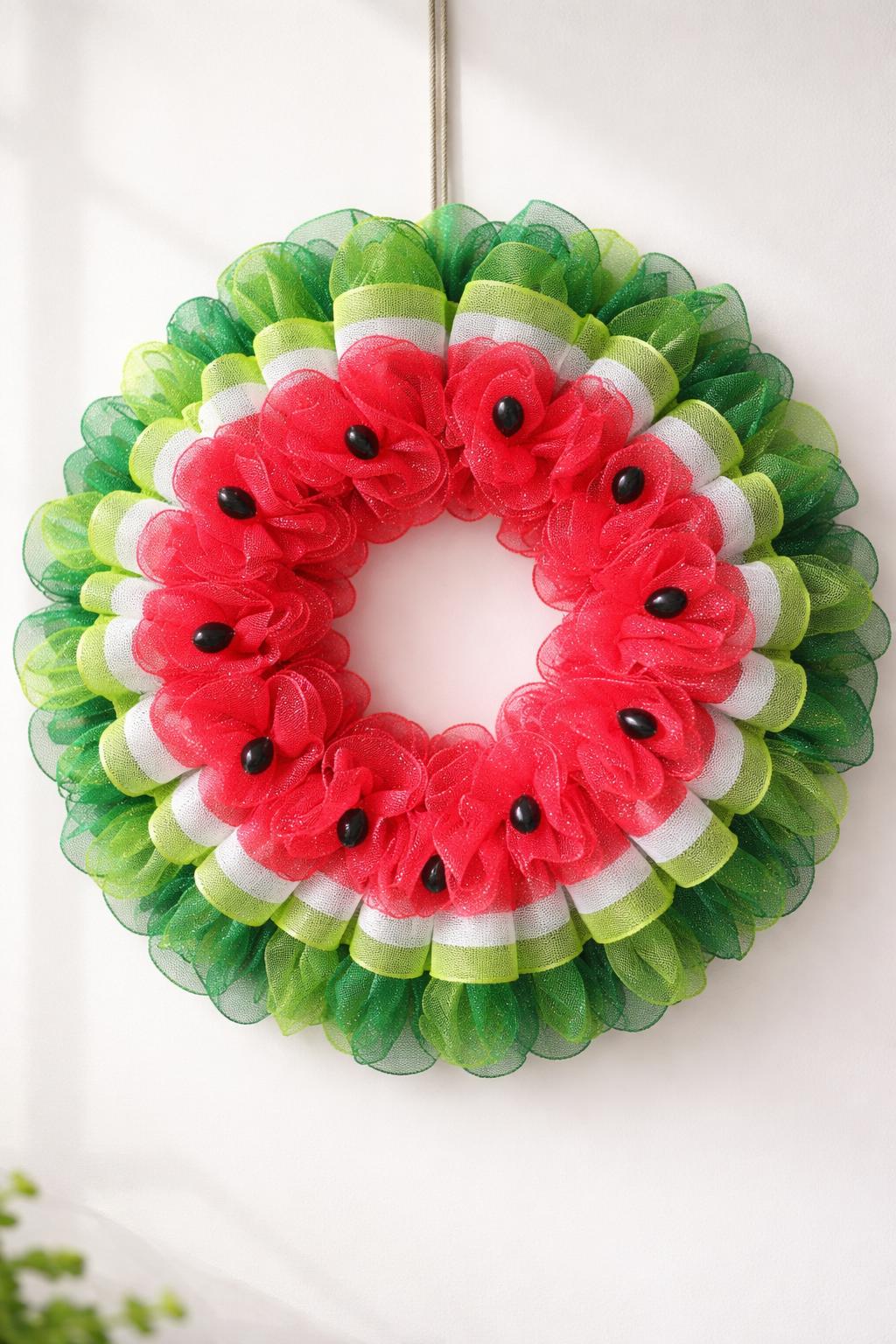 A colorful watermelon slice wreath made of mesh hanging against a plain background.