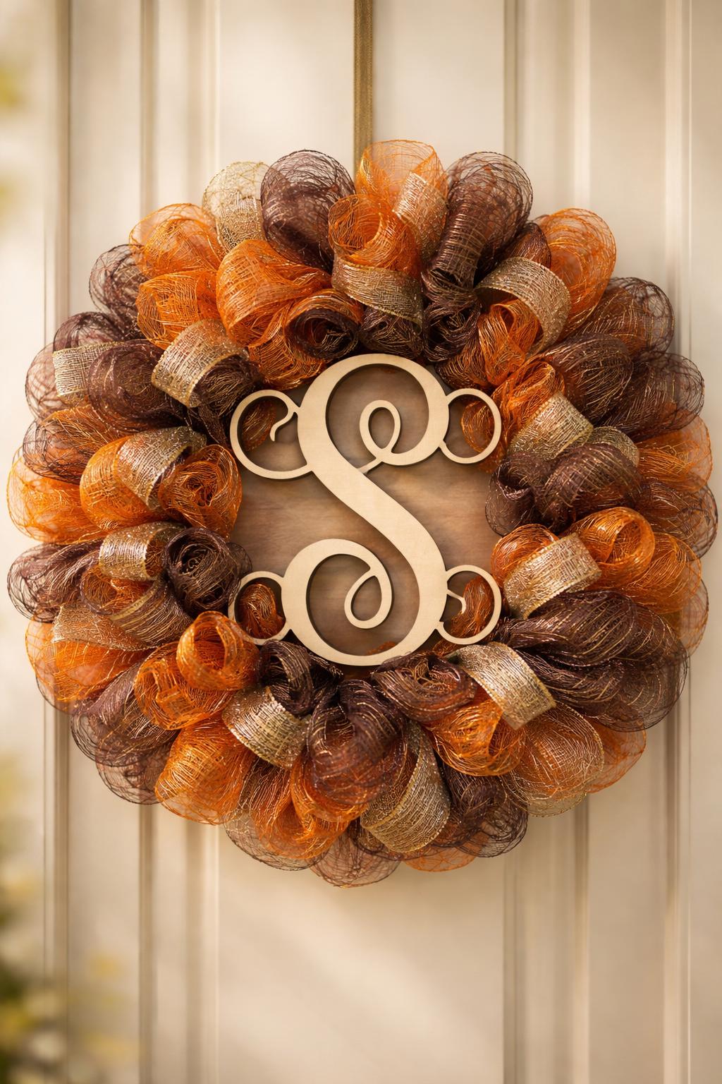 A circular wreath made of orange and brown mesh with a monogram centerpiece, displayed against a warm, neutral background.