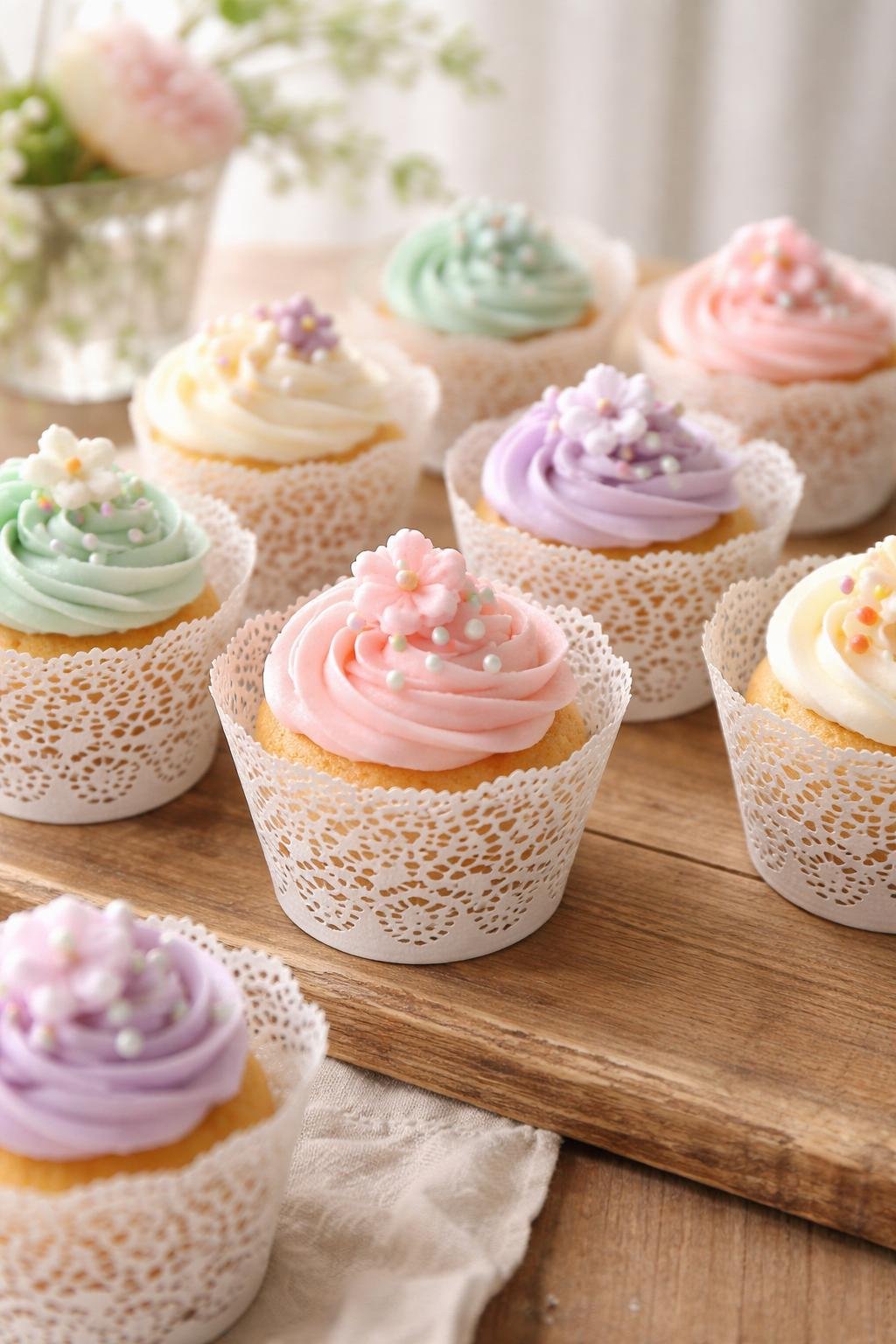 Cupcakes wrapped in white lace doily holders arranged on a wooden table with pastel-colored frosting and small decorative toppings.