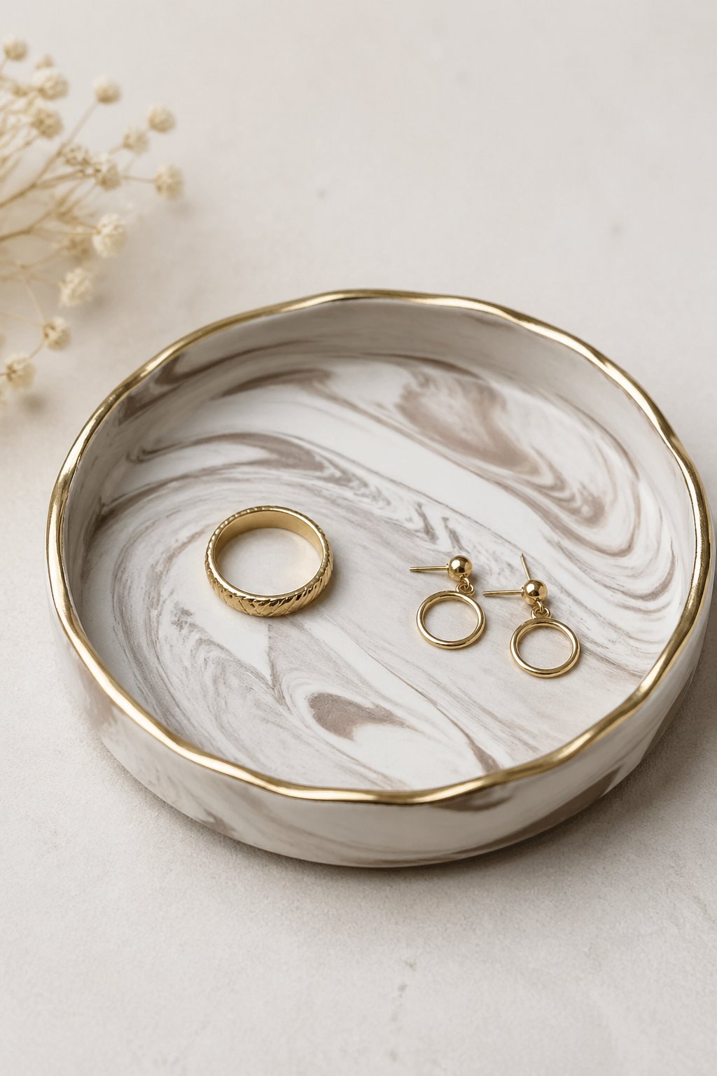 A marbled clay ring dish with a gold rim holding a gold ring and small earrings on a neutral background.