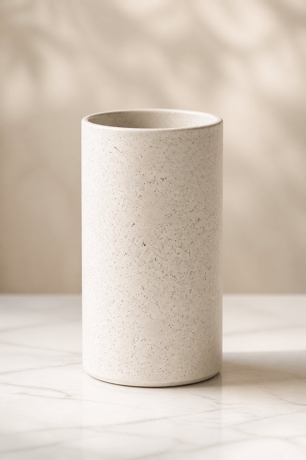 A simple cylinder vase with a speckled surface sitting on a light-colored surface against a neutral background.