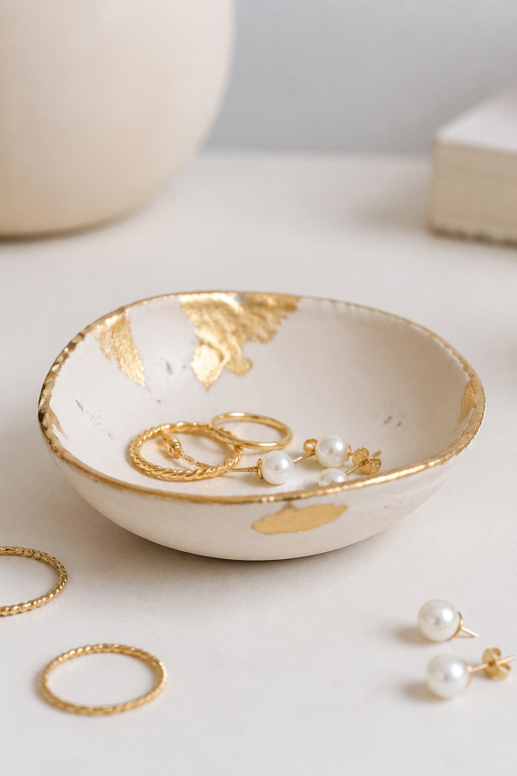 A small clay jewelry dish with gold leaf accents holding rings and earrings on a clean surface.