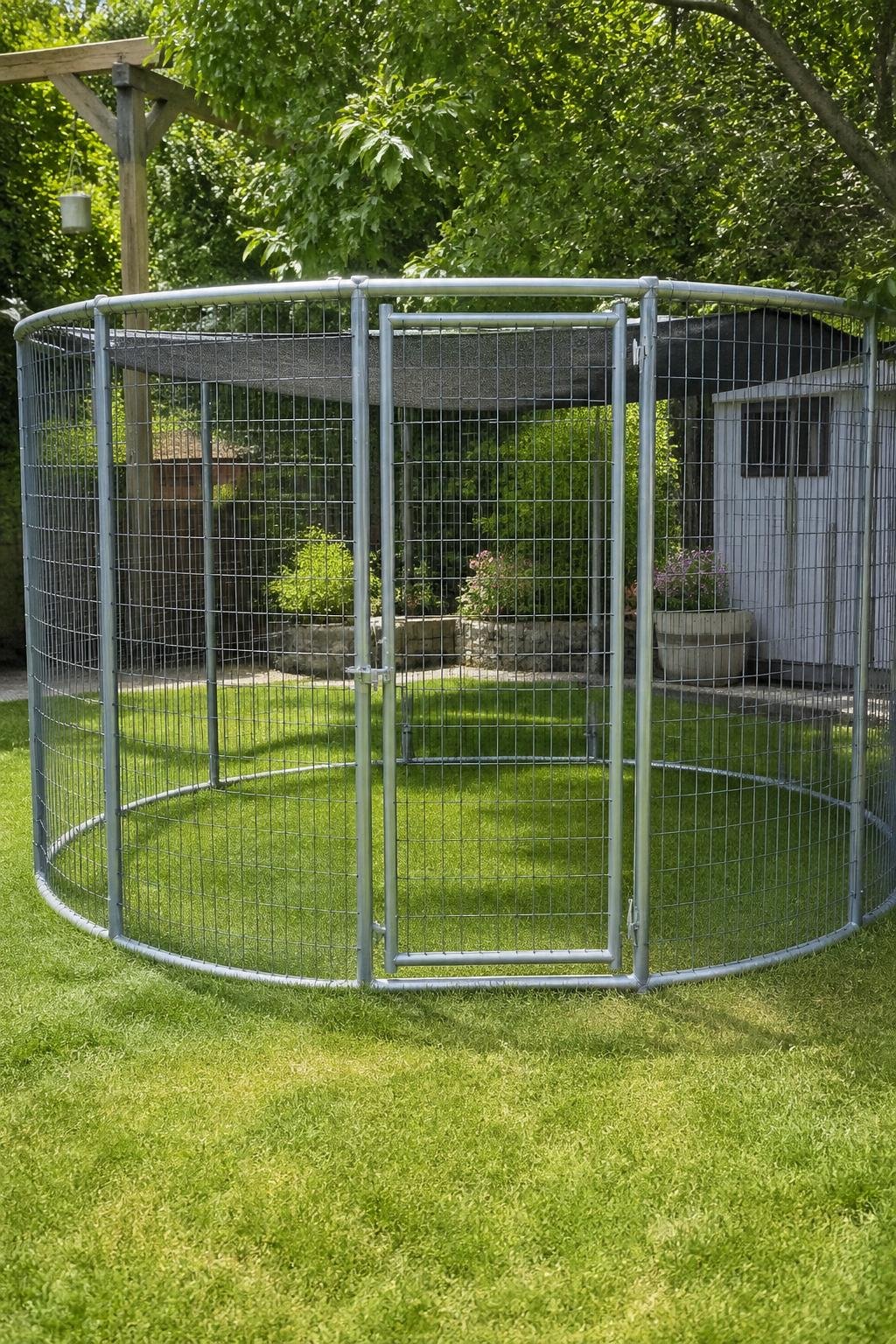 A backyard dog run made from a trampoline frame covered with welded wire mesh, set on green grass with plants around it.