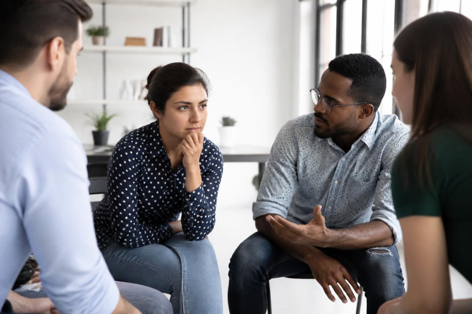 A group of adults in an office having a thoughtful discussion, with one woman looking contemplative and others listening attentively.