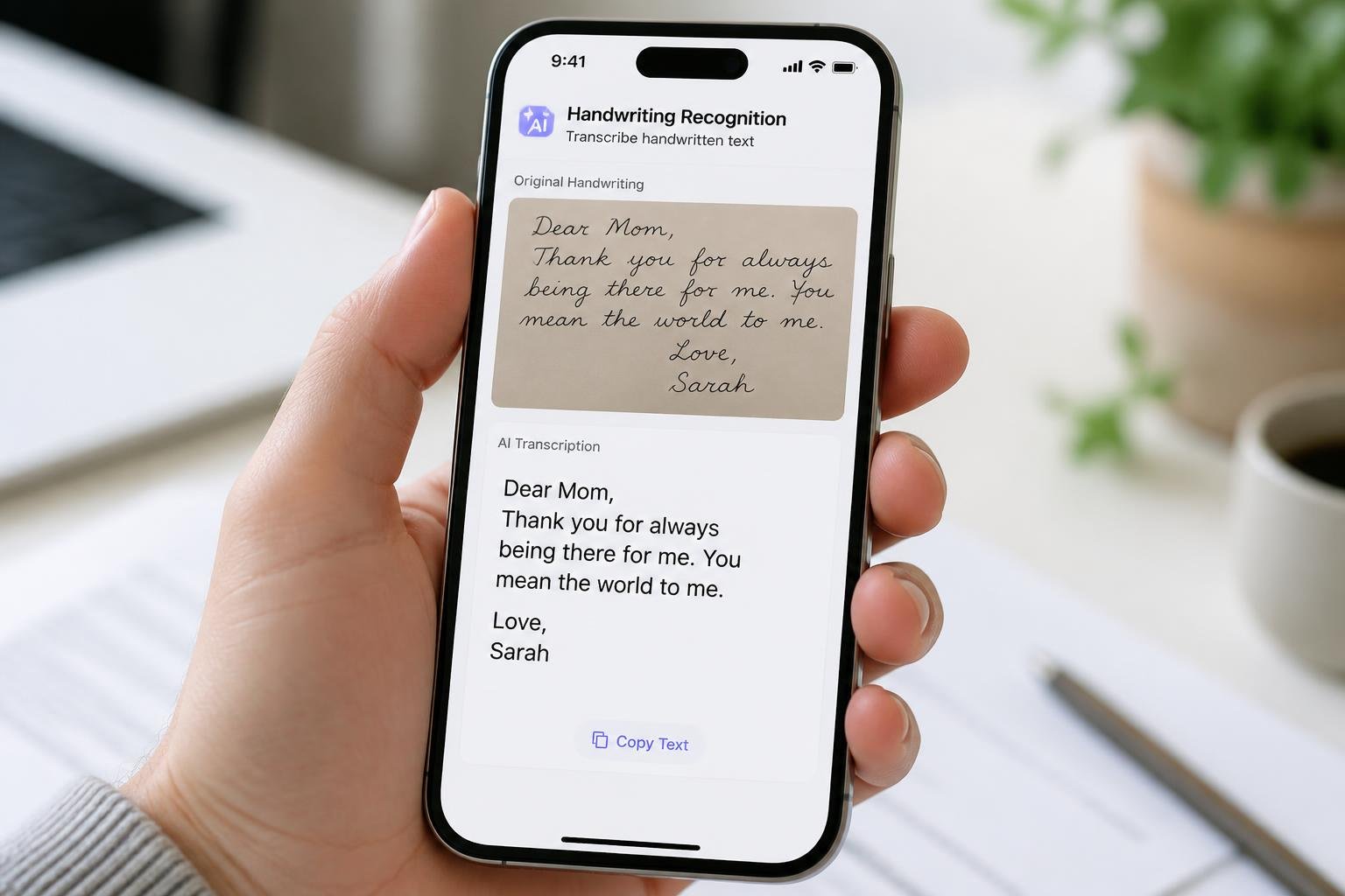 A hand holding an iPhone showing a close-up of cursive handwritten text on a card, with a blurred desk background.