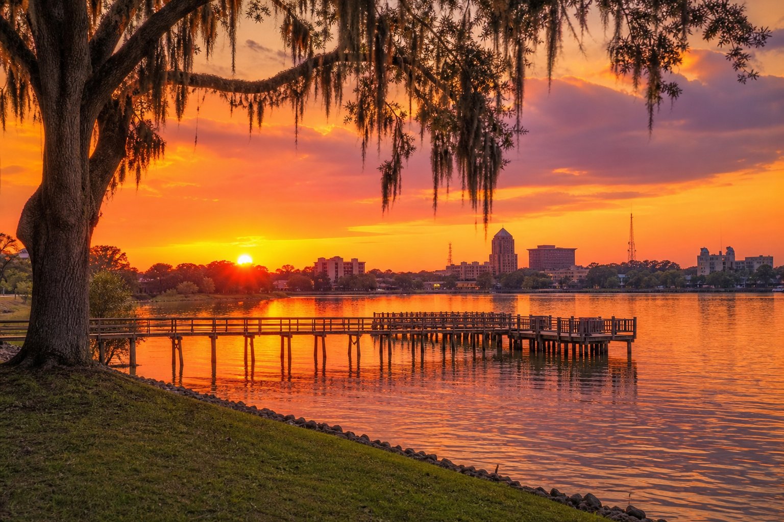 Sunset over a riverside park in Monroe, Louisiana, with oak trees, a wooden pier, and calm water reflecting colorful sky.