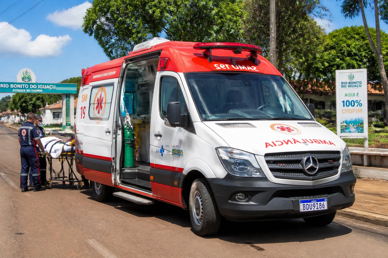 Ambulância estacionada em uma rua de Rio Bonito do Iguaçu com paramédicos próximos e árvores ao redor.