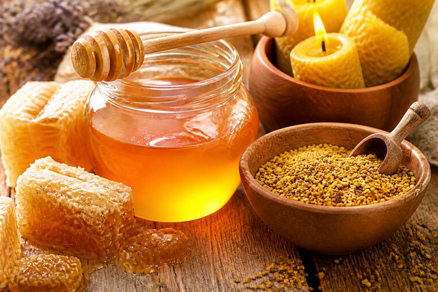 Close-up of honey jar, honeycomb, bee pollen, and beeswax candles arranged on a table.
