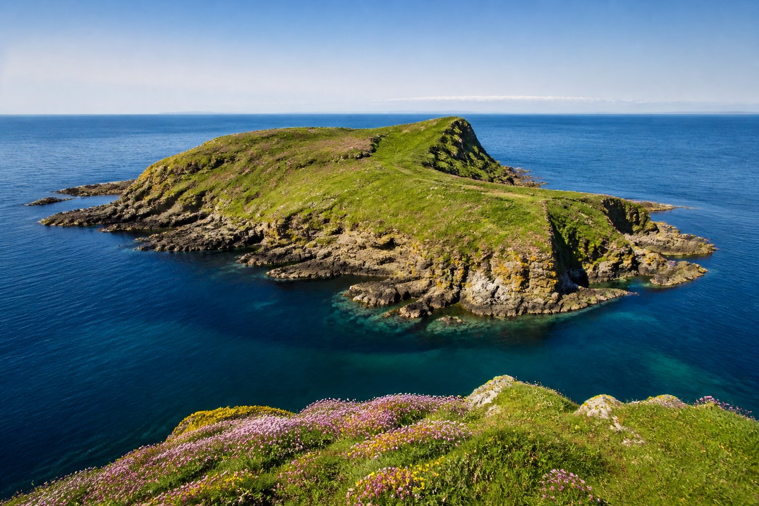 A small rocky island with green vegetation surrounded by calm blue water under a clear sky.