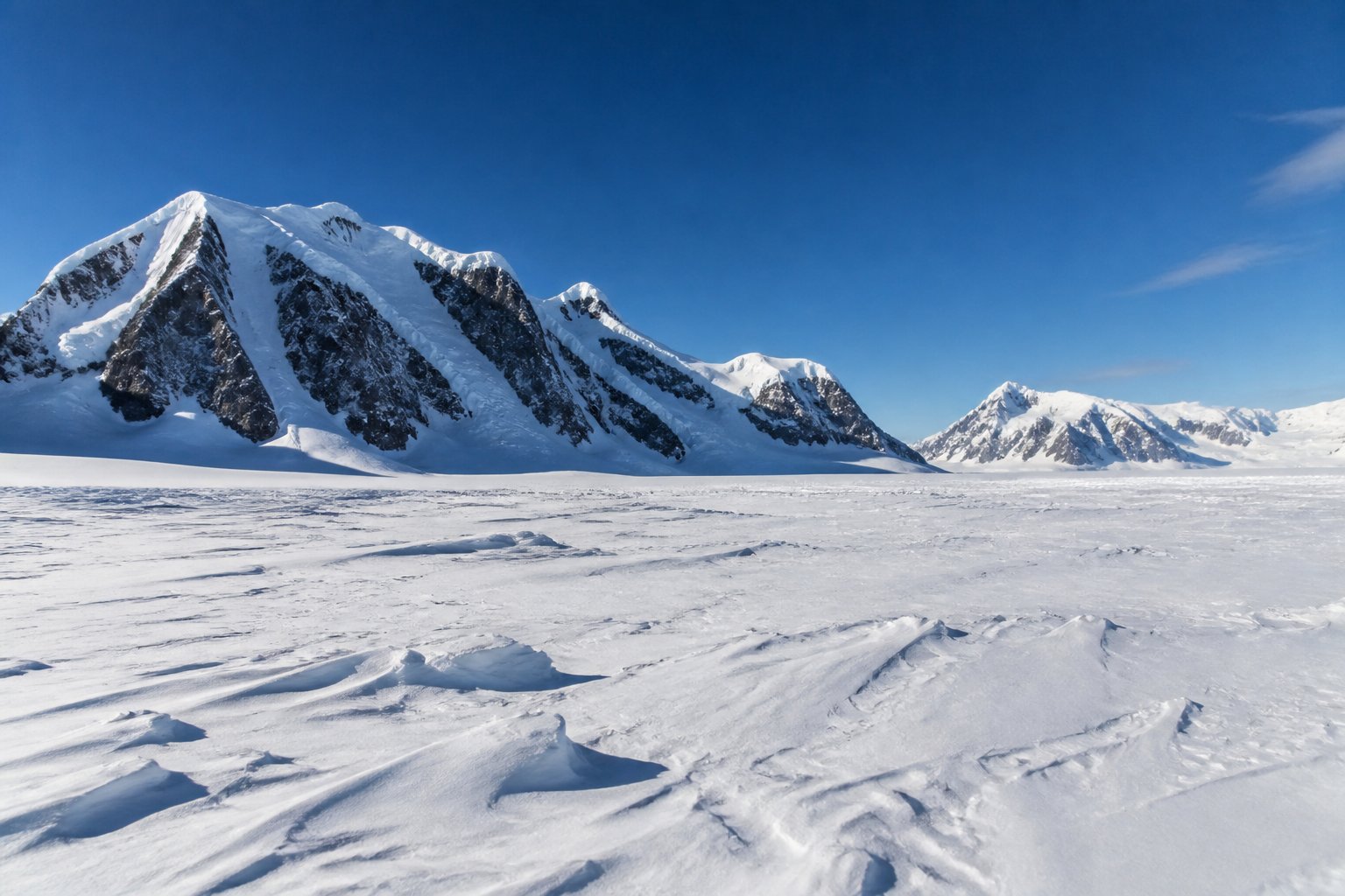 A snow-covered Antarctic landscape with mountains in the background and clear blue sky.