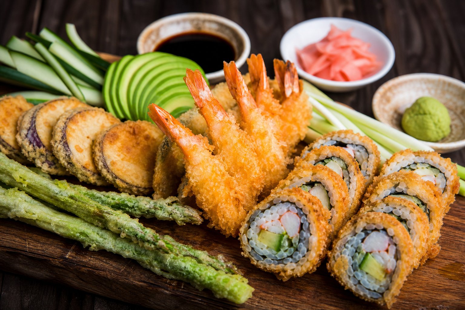 An assortment of fried sushi ingredients and fillings displayed on a wooden board with bowls of soy sauce and wasabi.