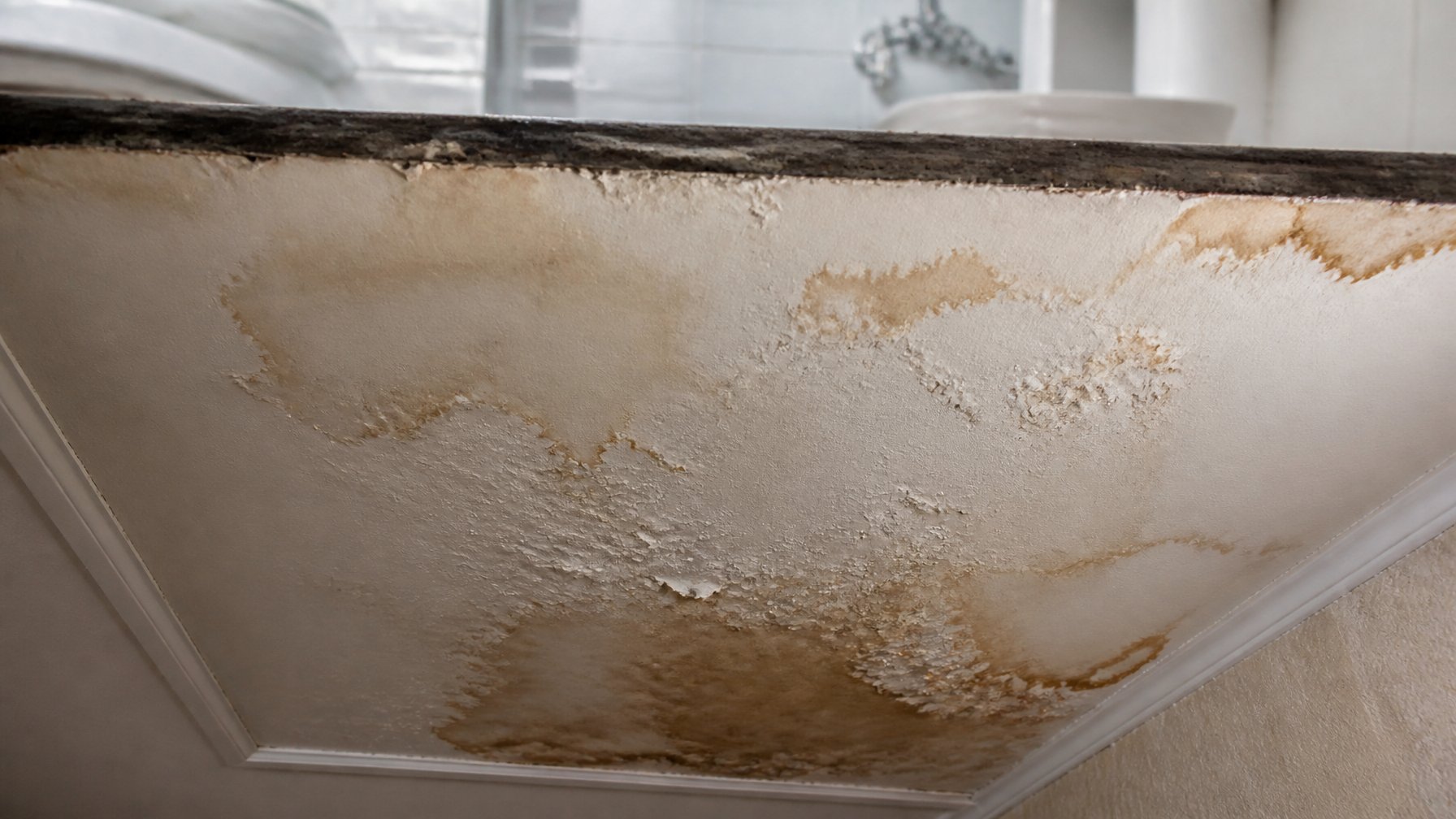 Close-up of a ceiling below a bathroom showing brown water stains and discoloration from water damage.
