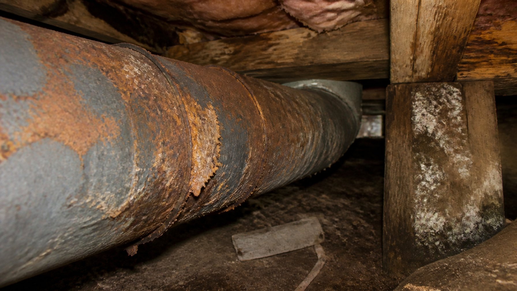 Close-up of rusted and corroded metal HVAC ductwork in a damp crawl space with wooden beams and exposed insulation.