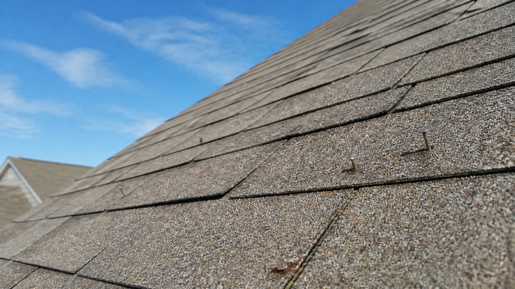 Close-up of a residential roof slope with missing or exposed nail heads on asphalt shingles under a clear sky.