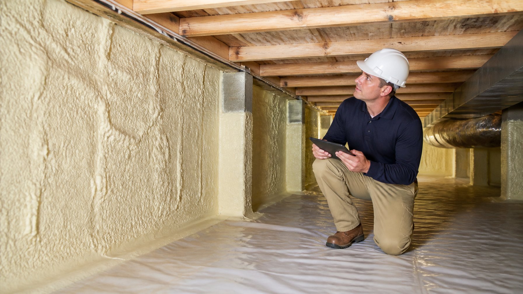 A person inspecting spray foam insulation in a clean, unvented crawl space beneath a house.