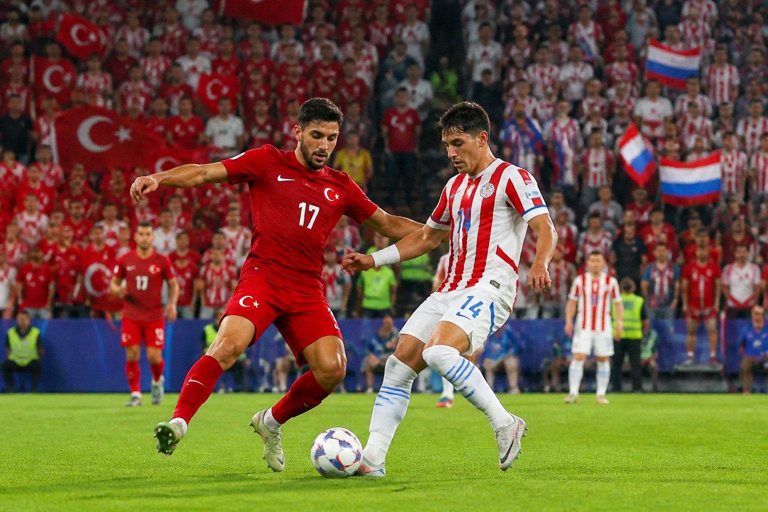 Türkiye ve Paraguay arasında oynanan bir Dünya Kupası futbol maçında sahada mücadele eden oyuncular ve tribünde tezahürat yapan taraftarlar.