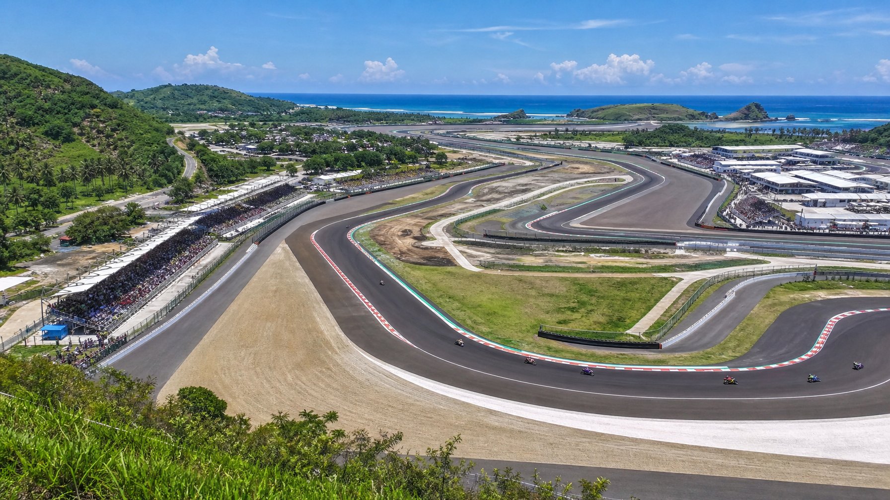Pemandangan udara sirkuit Mandalika dengan motor MotoGP sedang melaju di lintasan yang dikelilingi oleh pepohonan dan tribun penonton.