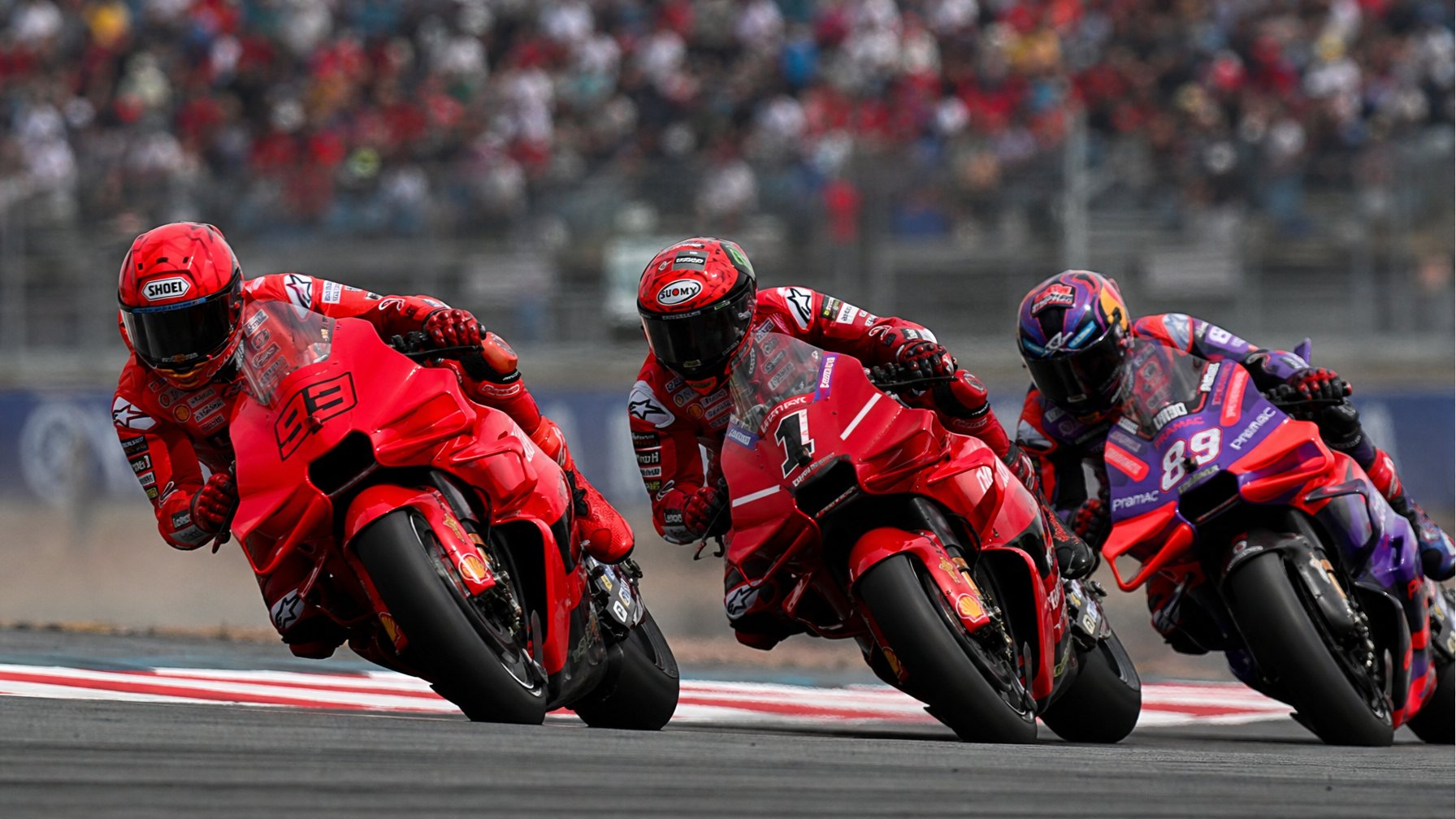 Tiga pembalap MotoGP sedang bersaing ketat di tikungan tajam pada lintasan balap, mengenakan perlengkapan balap lengkap dengan helm.