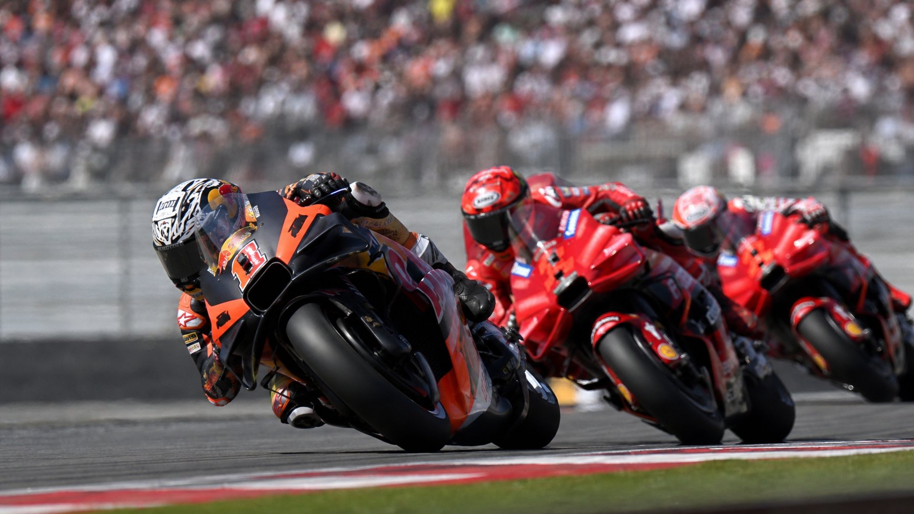 Seorang pembalap MotoGP sedang mengendarai motor dengan cepat di lintasan balap, dikelilingi oleh motor Ducati yang bersaing ketat.