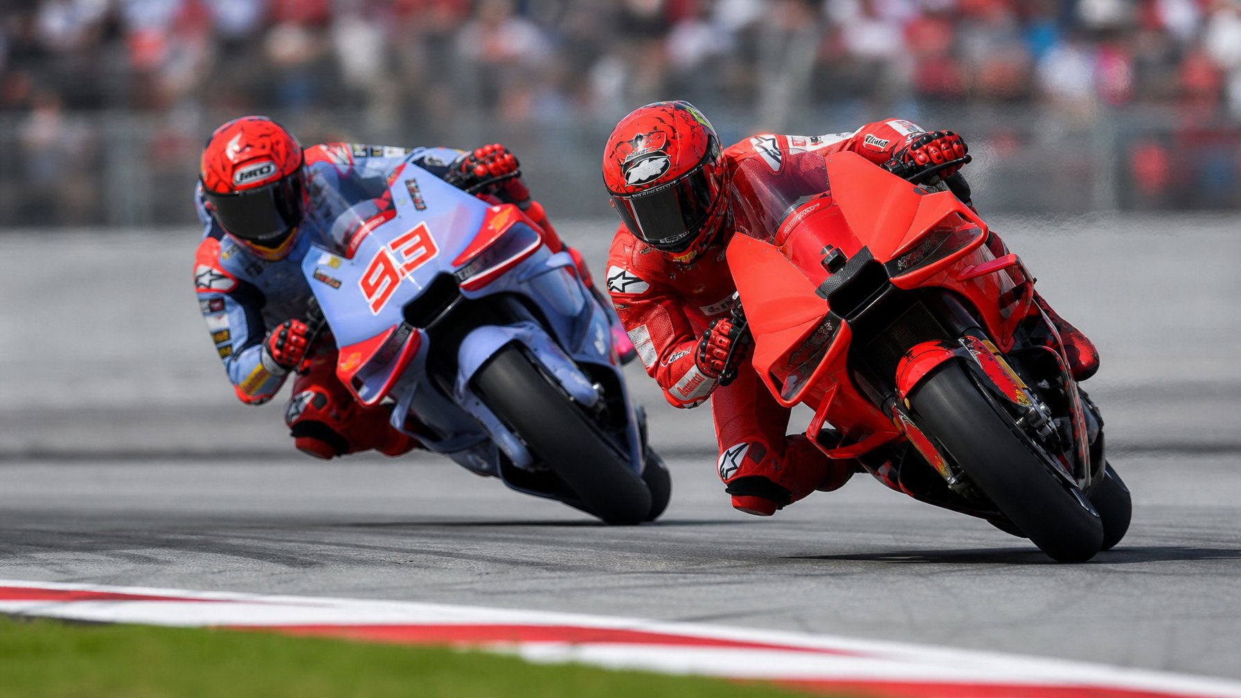 Dua pembalap MotoGP sedang bersaing ketat di tikungan lintasan balap dengan latar penonton dan suasana siang hari.
