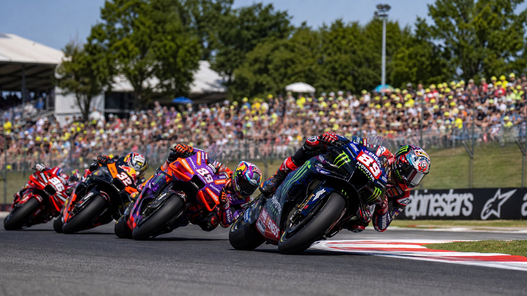 Balapan MotoGP dengan beberapa pembalap yang sedang menikung cepat di lintasan balap dikelilingi oleh penonton dan pepohonan.