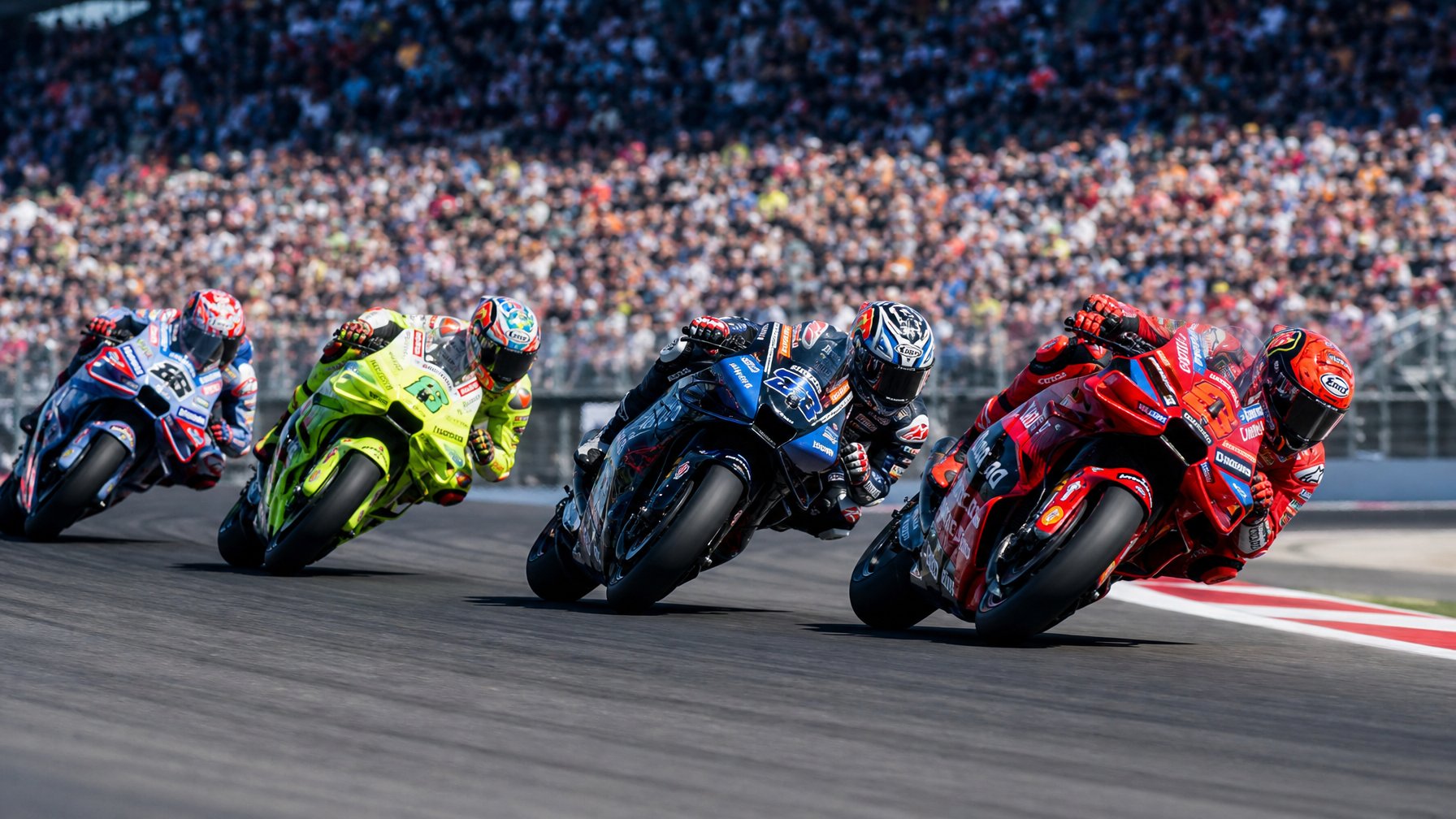 Beberapa pembalap MotoGP sedang balapan ketat di lintasan dengan penonton ramai di tribun.