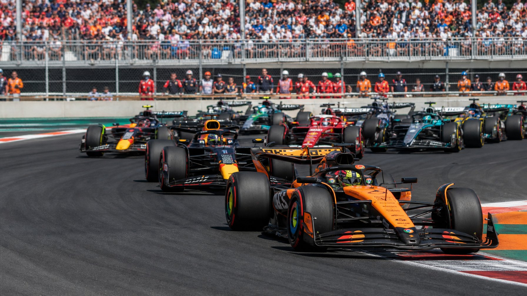 Beberapa mobil Formula 1 berwarna-warni melaju di trek balap dengan penonton dan kru tim di latar belakang.