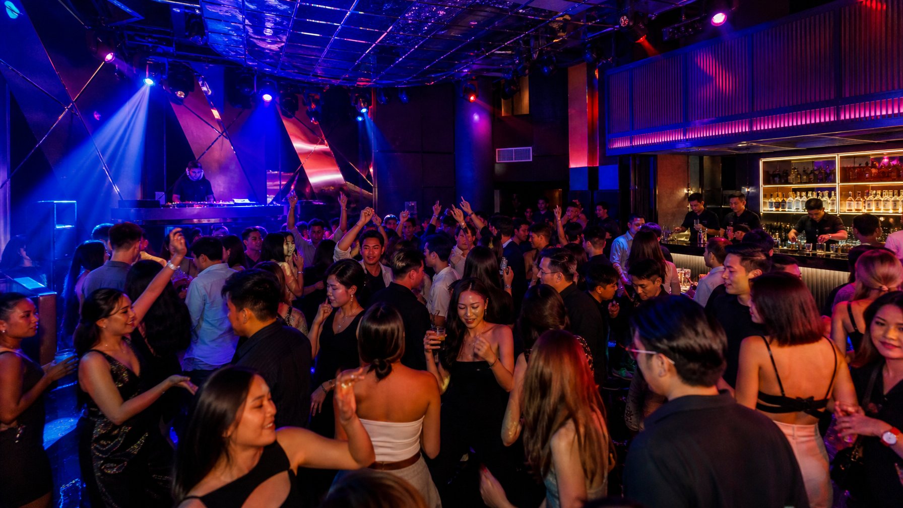 Orang-orang muda sedang berpesta dan menari di klub malam yang ramai dengan lampu warna-warni dan DJ yang sedang memutar musik.