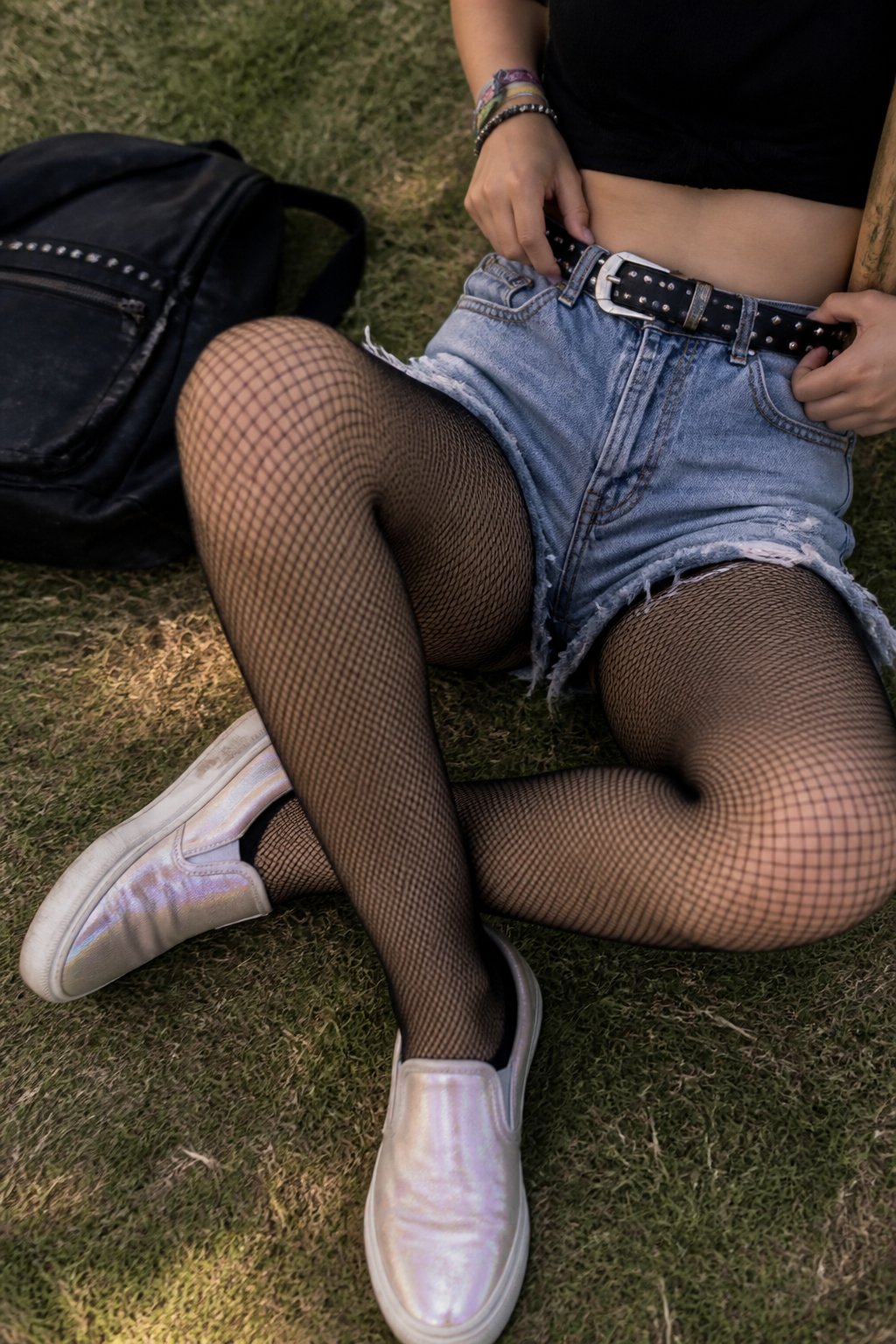 A person wearing pearlized slip-on Vans shoes, distressed shorts, fishnet tights, and a studded belt, photographed outdoors with natural daylight and casual surroundings.