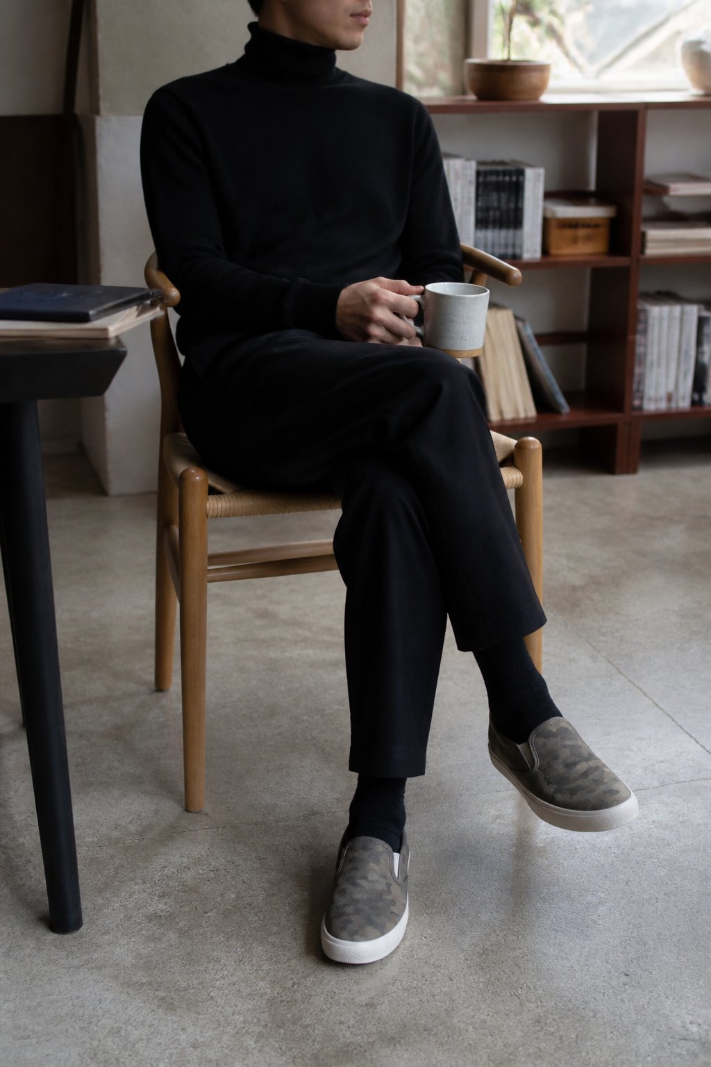 Person wearing pearlized camo slip-on Vans, black tapered trousers, and a fitted black turtleneck standing casually in soft daylight with natural shadows and a slightly off-center framing.