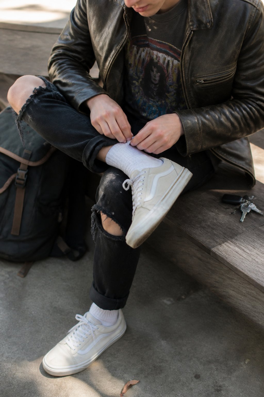 Person wearing pearlized Vans sneakers with ripped black jeans, a band t-shirt, and a leather jacket, sitting casually with hands adjusting the jacket, in soft natural daylight.