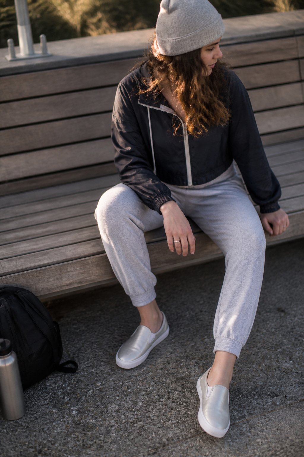 Person wearing pearlized slip-on Vans with joggers, a cropped windbreaker, and a beanie, standing casually outdoors with natural daylight and everyday objects nearby.