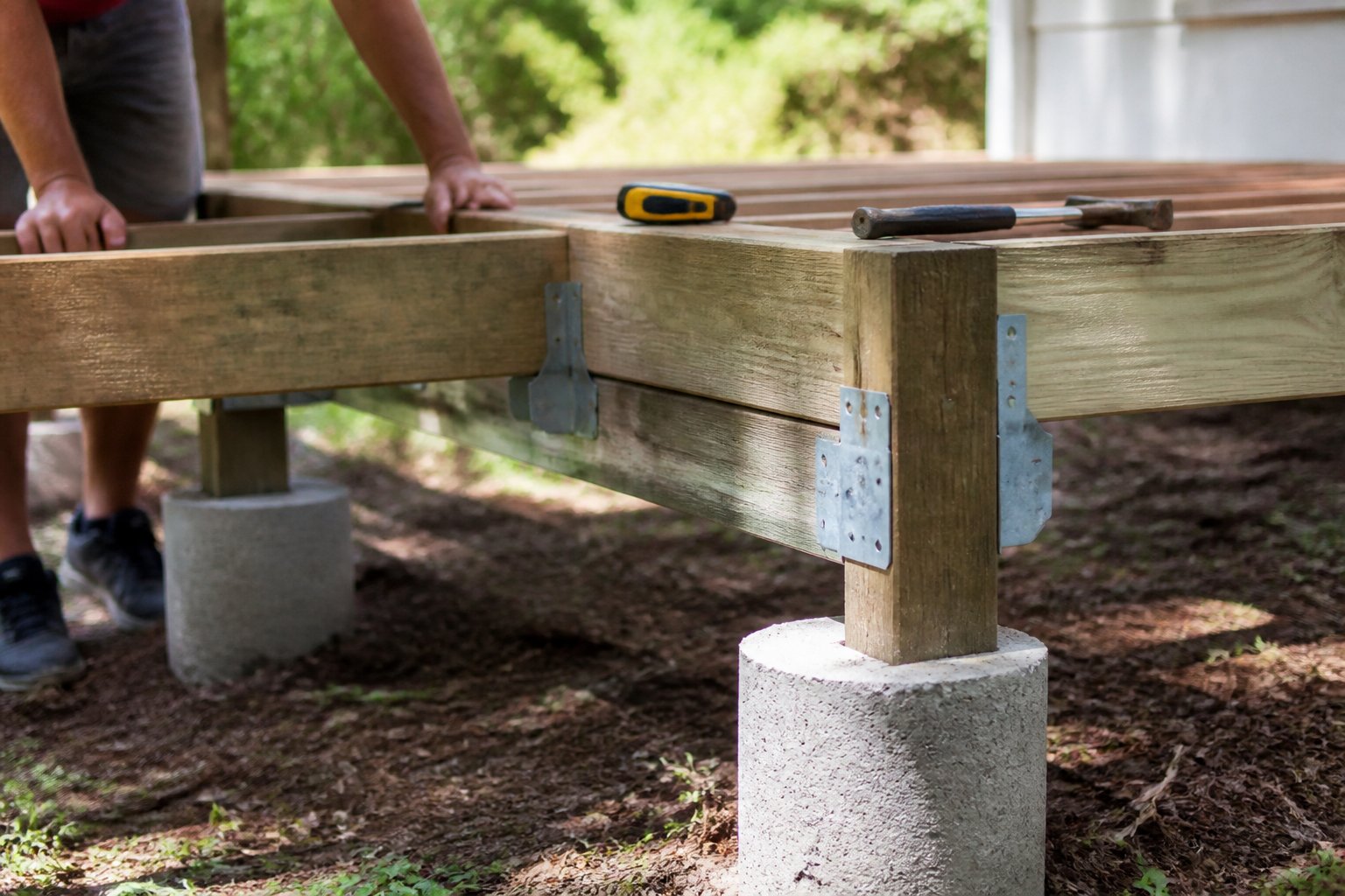 A simple raised wooden deck built on concrete piers with galvanized joist hangers, with tools and hands working on it in natural daylight.