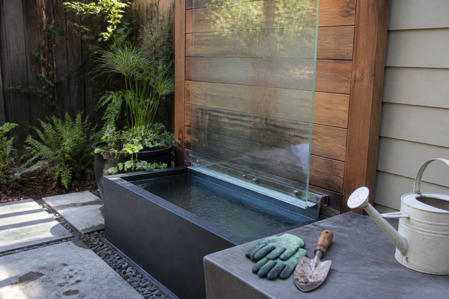Backyard water feature with a tempered glass panel and cedar wood wall over a water basin surrounded by gardening tools and gloves.