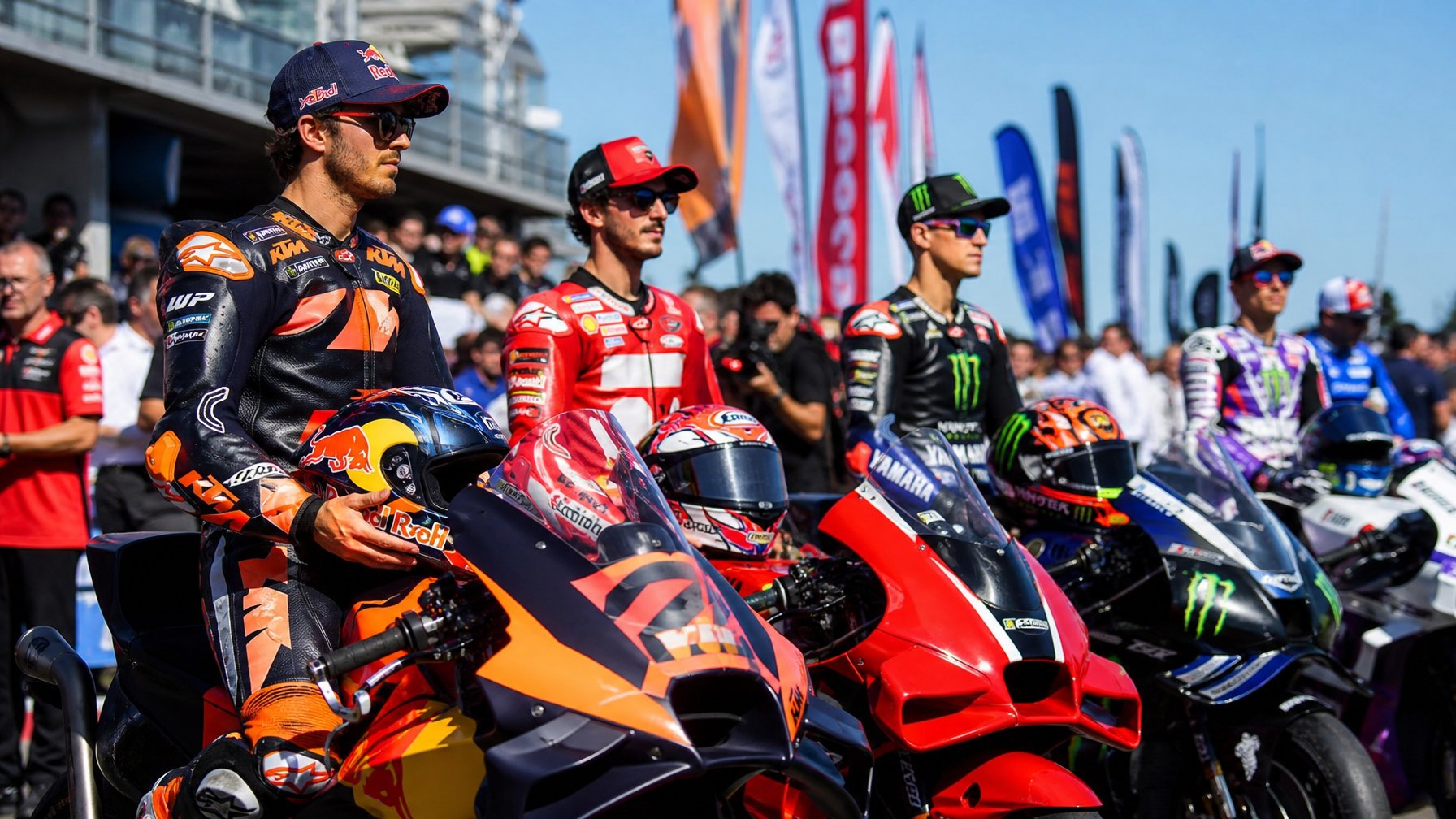 Beberapa pembalap MotoGP berdiri di samping motor balap mereka di area paddock dengan tim dan media di latar belakang.