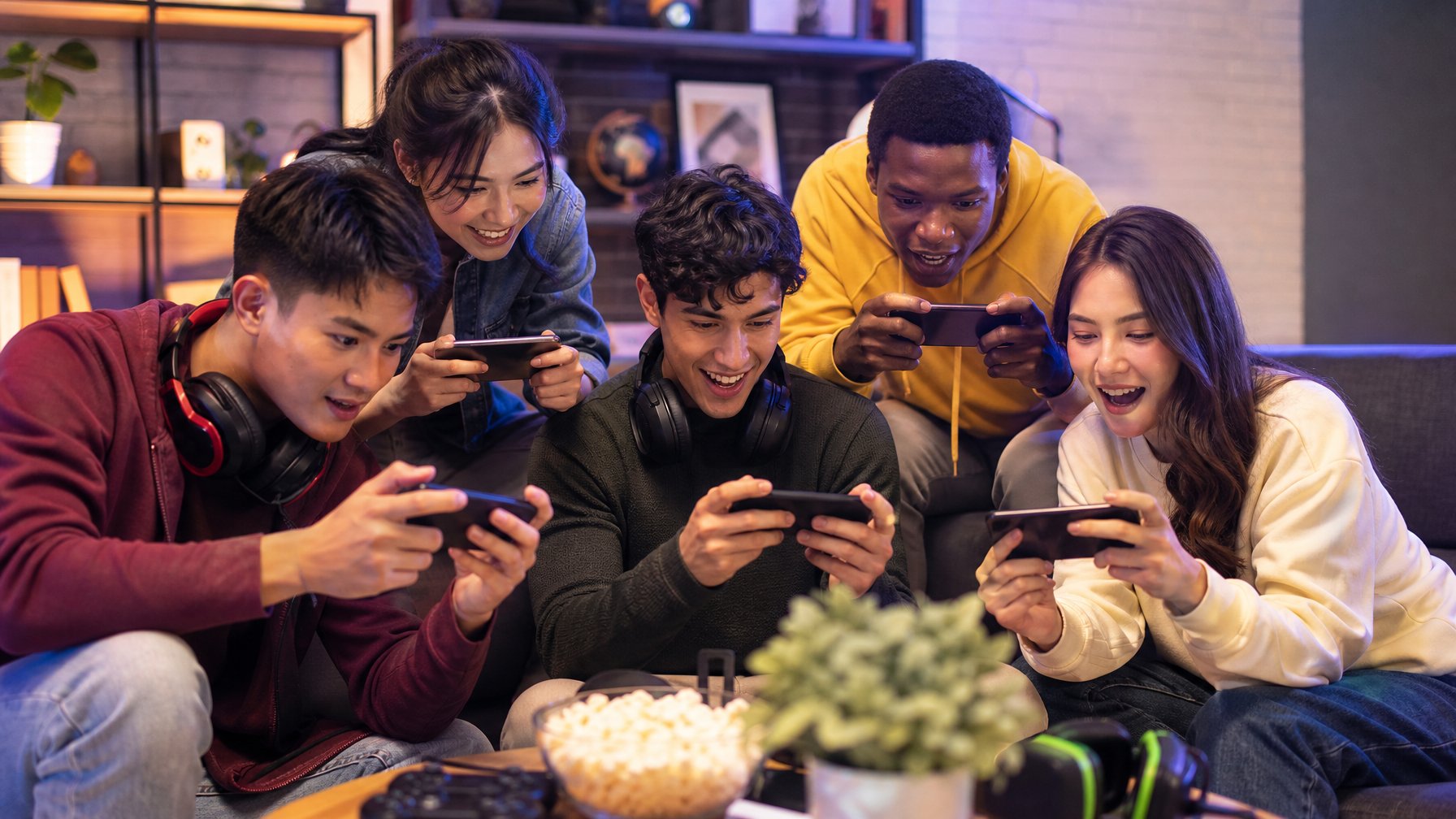 Sekelompok orang muda bermain game mobile bersama di ruang tamu yang nyaman, tampak fokus dan bersemangat.