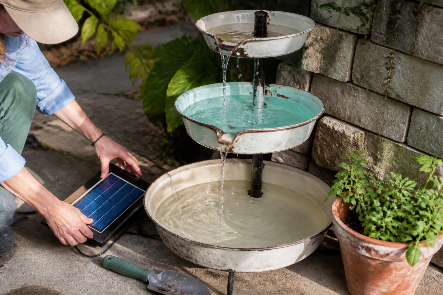 A vintage tiered wash basin fountain with a solar pump outdoors, surrounded by gardening tools and a small potted plant, with hands adjusting the pump.