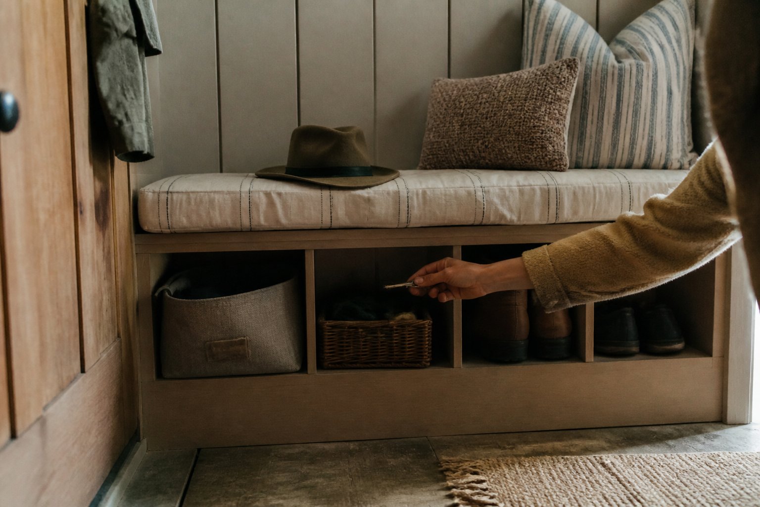 A compact mudroom bench with cubby storage holding shoes and personal items, with a hand reaching in, bathed in soft natural daylight.