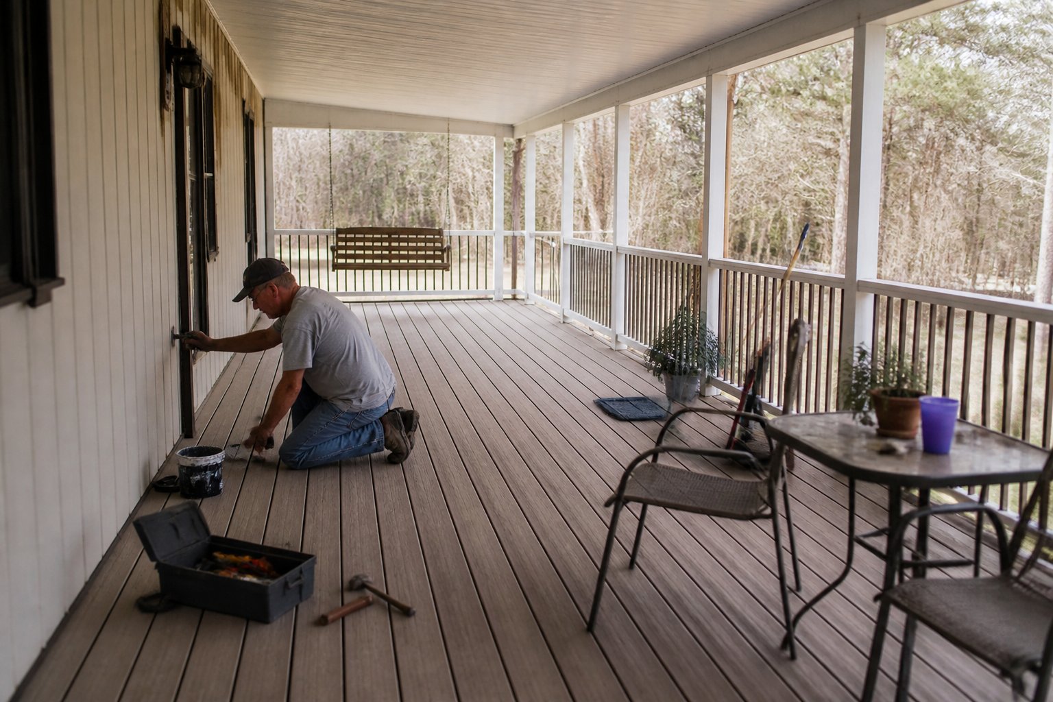 A covered porch with composite decking and railings attached to a manufactured home, with hands holding tools and a small toolbox on the porch floor.