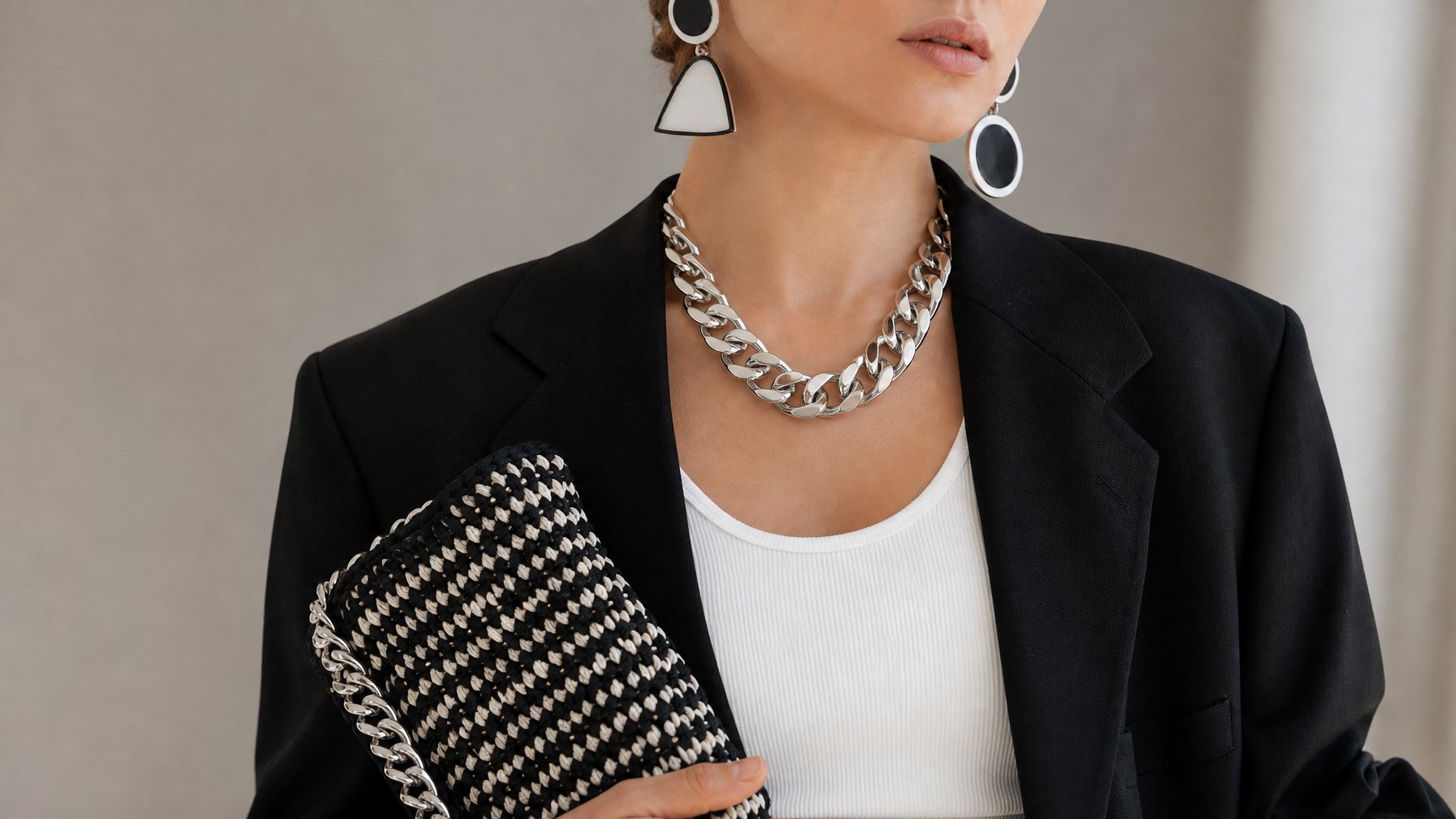 Seorang model mengenakan pakaian hitam putih dengan aksesori besar yang mencolok, termasuk anting-anting geometris dan kalung perak, di latar belakang minimalis.