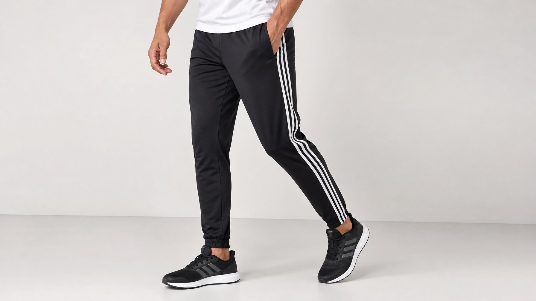 Seorang pria mengenakan celana training Adidas hitam dengan garis putih di samping, berdiri dengan latar belakang polos.