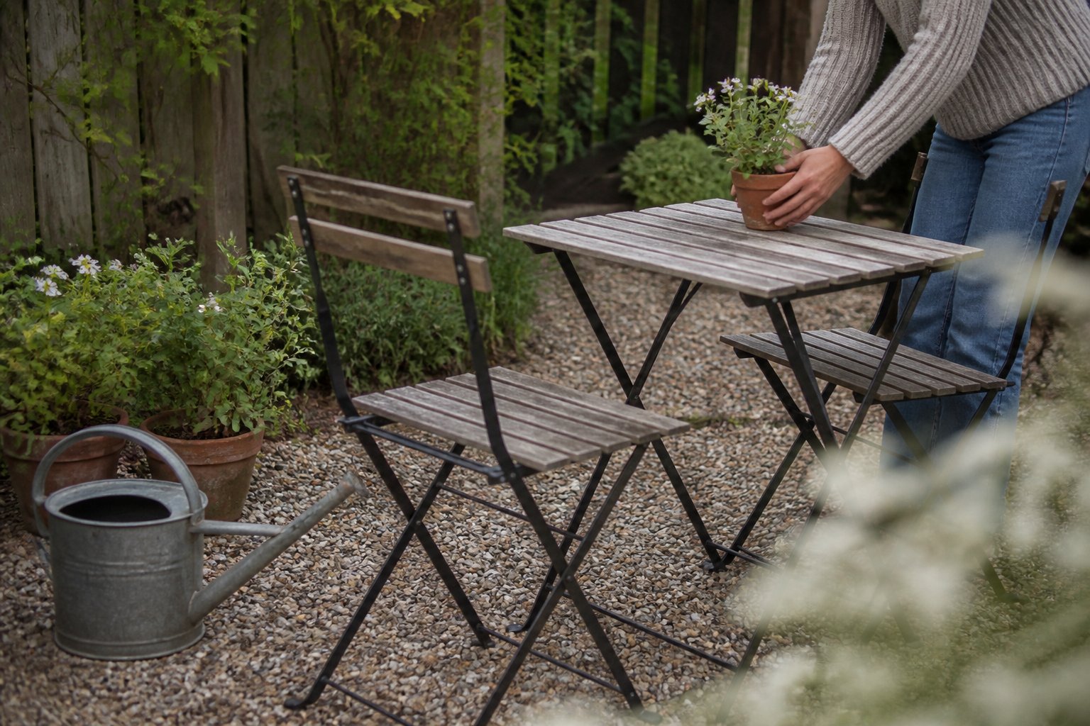 A pea-gravel patio with a bistro set surrounded by gardening tools and a potted plant, bathed in soft daylight.