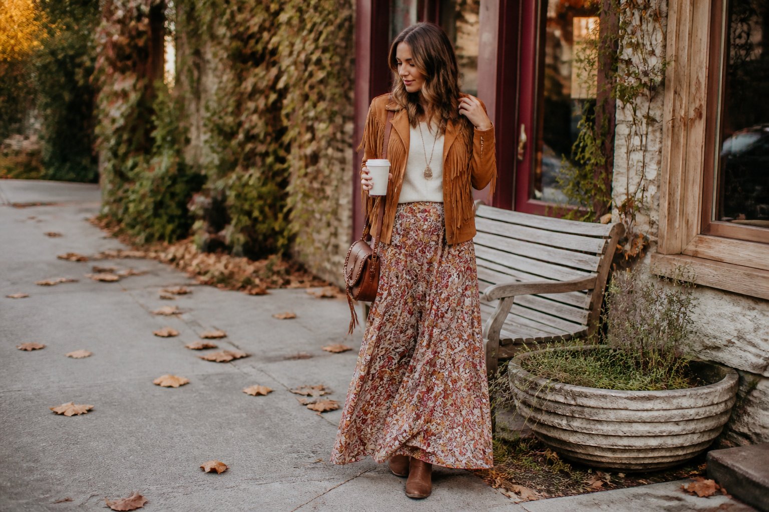 A woman wears a tan fringe jacket with a floral maxi skirt on a leafy autumn sidewalk.