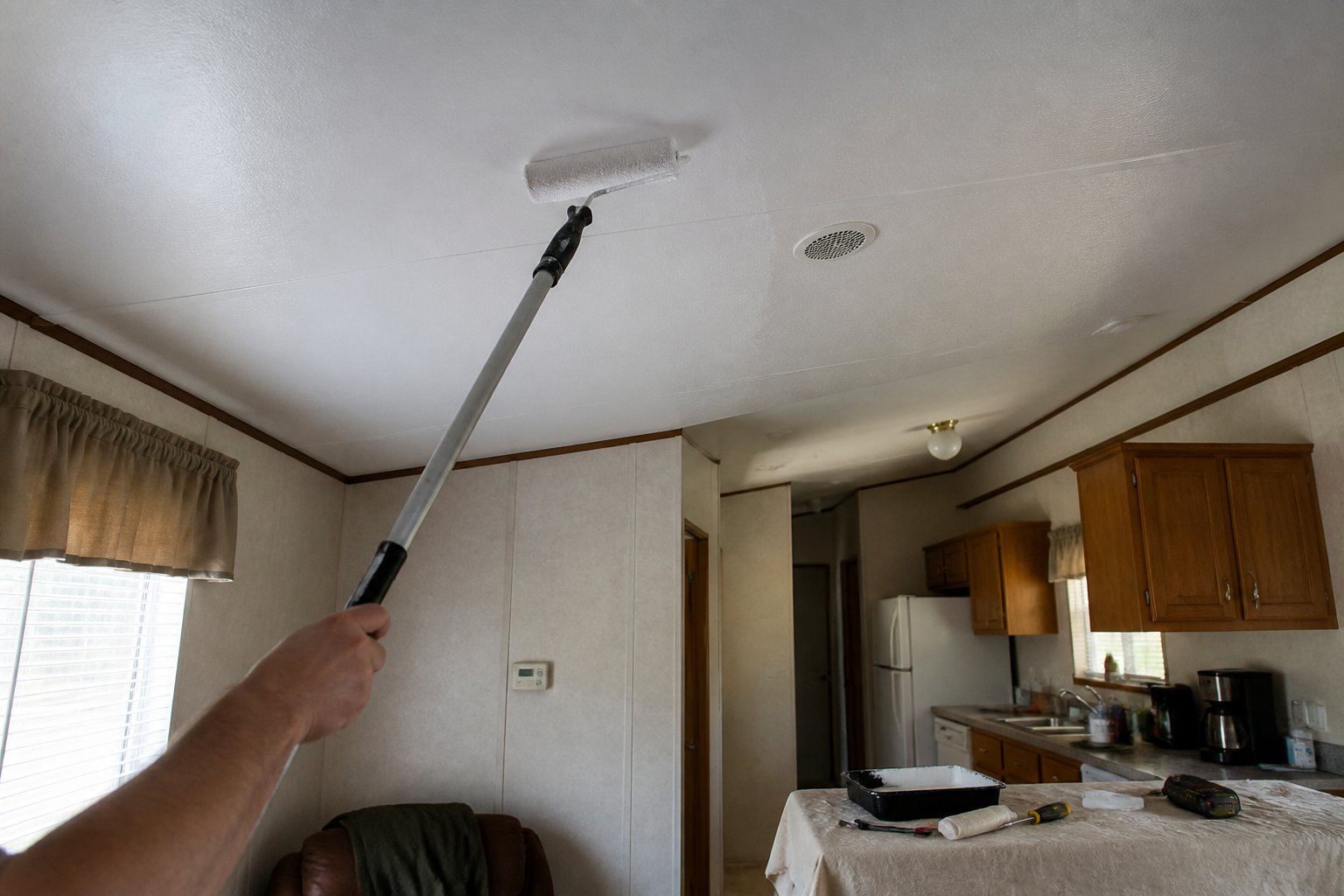 A homeowner paints a bright white ceiling in a tidy mobile home while a paint tray and tools sit nearby.