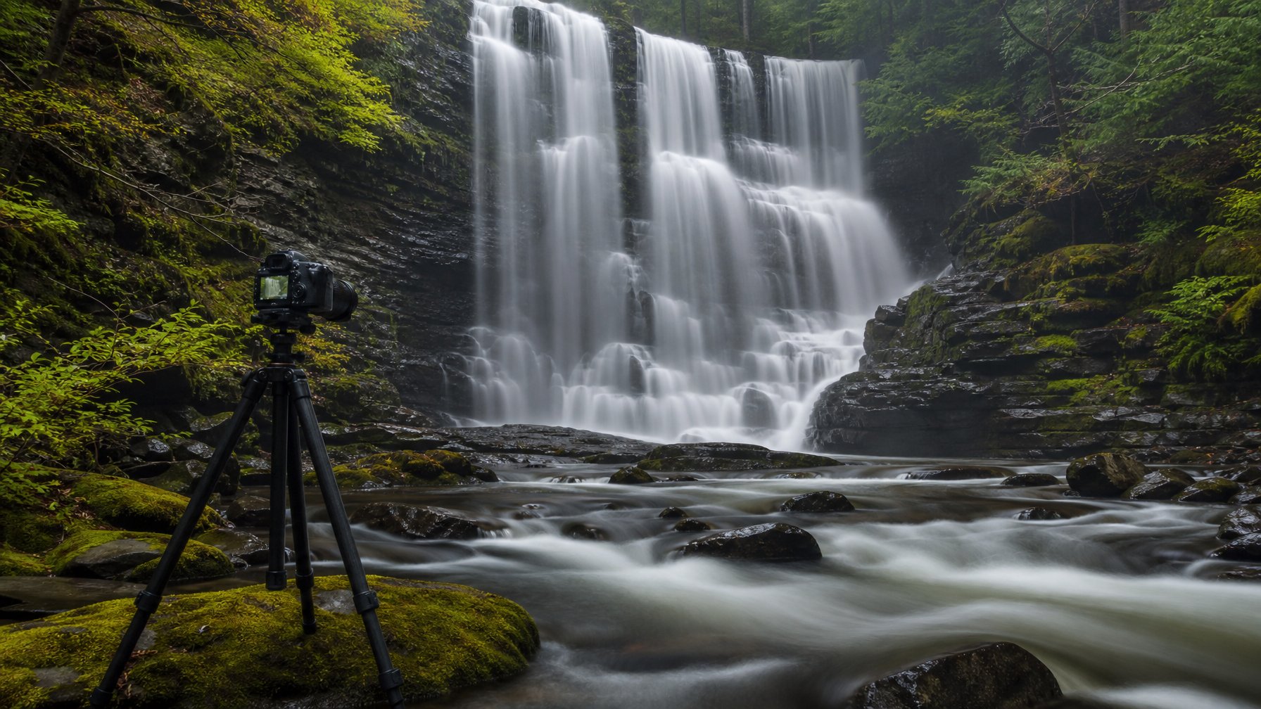 Air terjun mengalir lembut ke sungai di tengah hutan hijau dengan kamera di atas tripod.