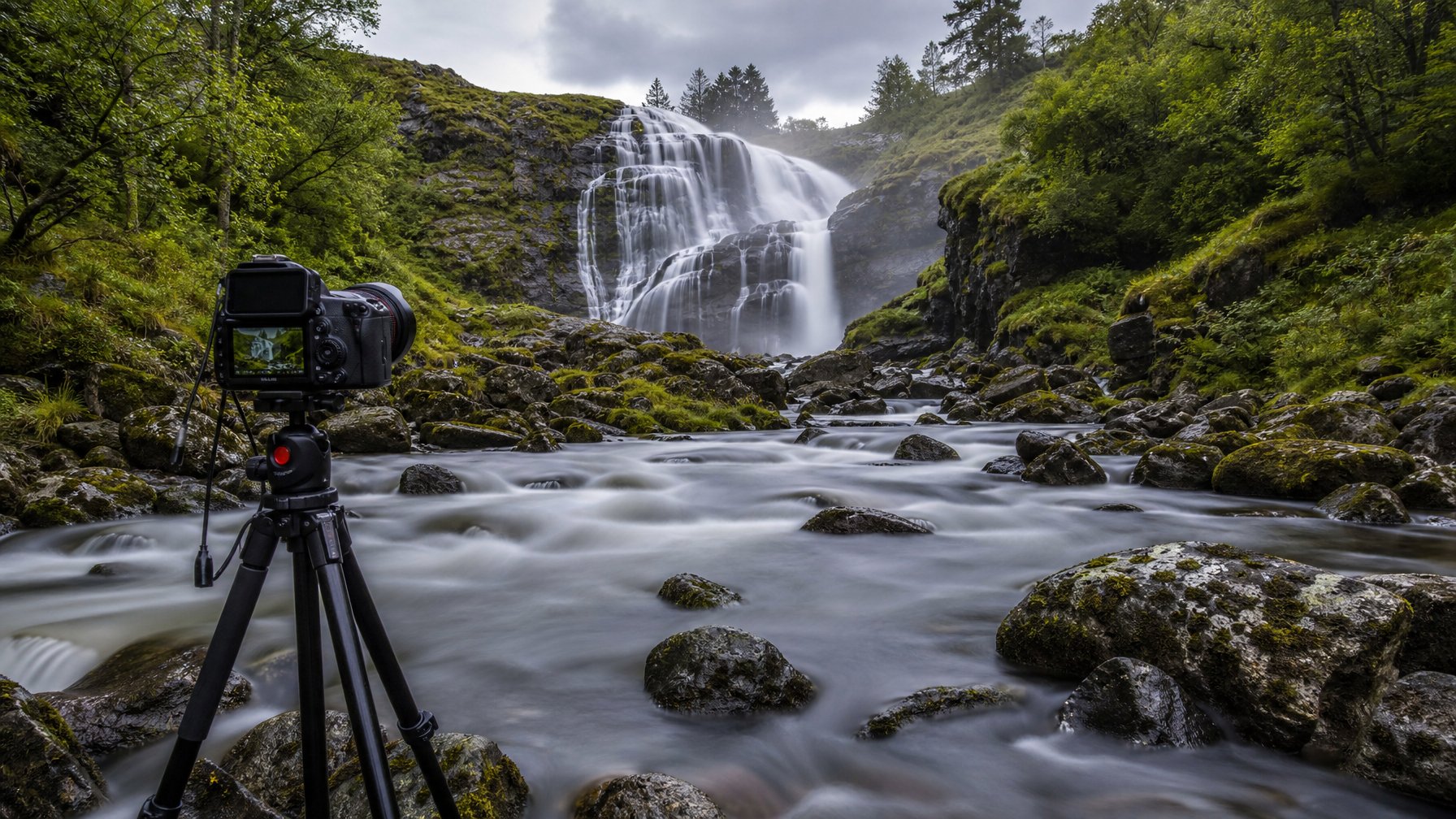 Kamera pada tripod menghadap air terjun dan sungai dengan aliran air yang tampak lembut.